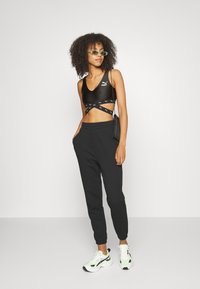 Sujeta deportiva negra con detalle cruzado y logo, combinada con pantalones de chándal negros que tienen bolsillos laterales y puños elásticos, complementada con zapatillas.