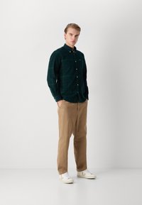 Camisa xadrez verde com botões, tecido de algodão, mangas longas. Calças bege, leves. Ténis brancos com logótipo dos lados. Look simples e casual.