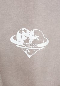 Il tessuto beige presenta un design grafico bianco di un globo circondato da un cuore e il testo "SMILODOX" in un ovale sottostante.