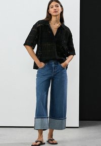 Schwarzes Spitzenhemd mit kurzen Ärmeln, kombiniert mit hochgeschnittenen, weit geschnittenen blauen Jeans mit fransendem Saum. Schwarze Flip-Flops runden den Look ab.