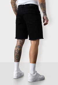 Black denim shorts with frayed hem, back pockets, worn with white sneakers and white socks. Displaying detailed tattoos on the left leg.