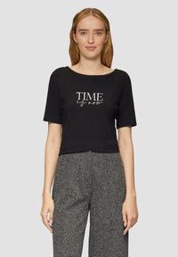 Schwarzes, kurzes T-Shirt aus Baumwolle mit kurzen Ärmeln. Verziert mit dem Spruch "TIME is now" in metallischer Schrift. Das Model trägt graue gemusterte Hosen.