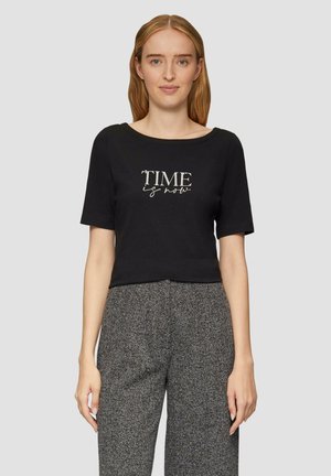 T-shirt noir court en coton avec des manches courtes. Comprend la phrase "LE TEMPS c'est maintenant" en impression métallique. Le modèle porte un pantalon à motifs gris.