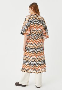 Długi kimono o wzorze zygzakowym w kolorach pomarańczowym, czarnym i białym. Posiada szerokie rękawy i pasek do związania w talii. Prezentowane na jasnych spodniach.