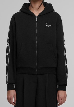 Personne portant un sweat à capuche noir zippé avec texte blanc et motifs d'étoiles le long des manches sur un fond gris uni.
