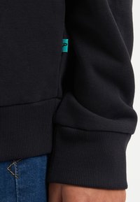 Tejido de sudadera negra con textura acanalada, que presenta una etiqueta turquesa visible y un detalle de puño ajustado.