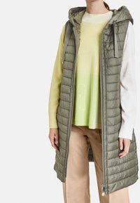 Vadderad olivgrön väst med huva, utrustad med hel dragkedja framtill, texturerad quiltad design och sidofickor, lager-på-lager över en lätt tröja.