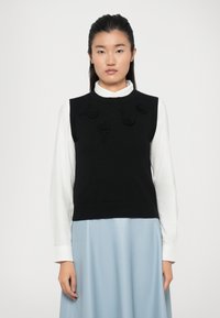 Un pull sans manches en tricot noir avec des appliqués floraux à l'avant, porté sur une chemise blanche à col, associé à une jupe bleu clair.