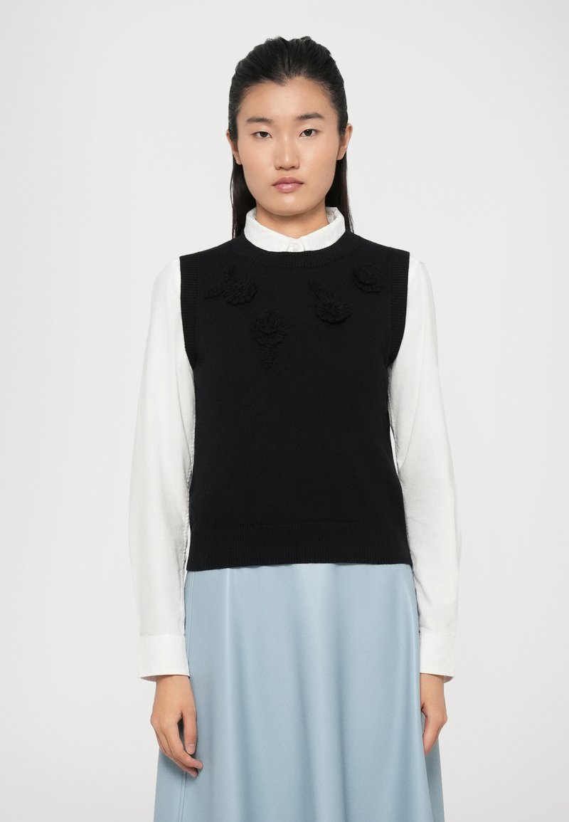 Un pull sans manches en tricot noir avec des appliqués floraux à l'avant, porté sur une chemise blanche à col, associé à une jupe bleu clair.