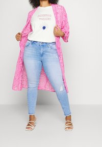 En person poserar i ljusblå jeans och en grafisk T-shirt under en rosa mönstrad kimono, iförd vita sandaletter med remmar.