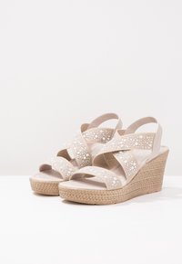 Sandales compensées beige avec des brides croisées, ornées de perles blanches décoratives, et une semelle plateforme texturée enveloppée de jute.