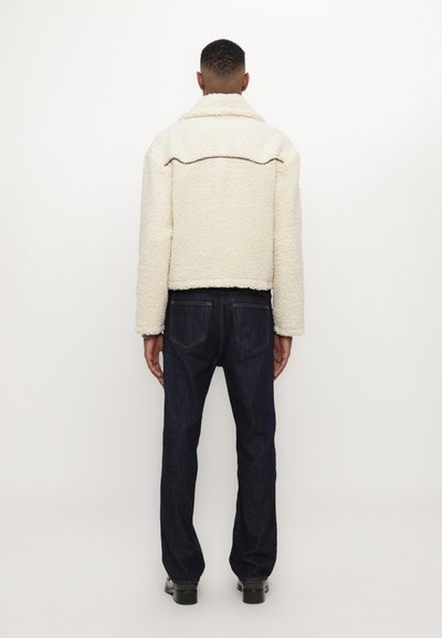 Cream-colored, textured fleece jacket with a collar, back seam detail, paired with dark blue denim jeans and black footwear.