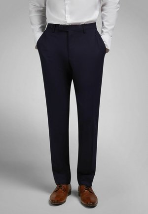 Mann trägt marineblaue Anzughose, weißes Hemd eingesteckt, Hände in den Taschen und braune Lederschuhe vor schlichtem grauem Hintergrund.