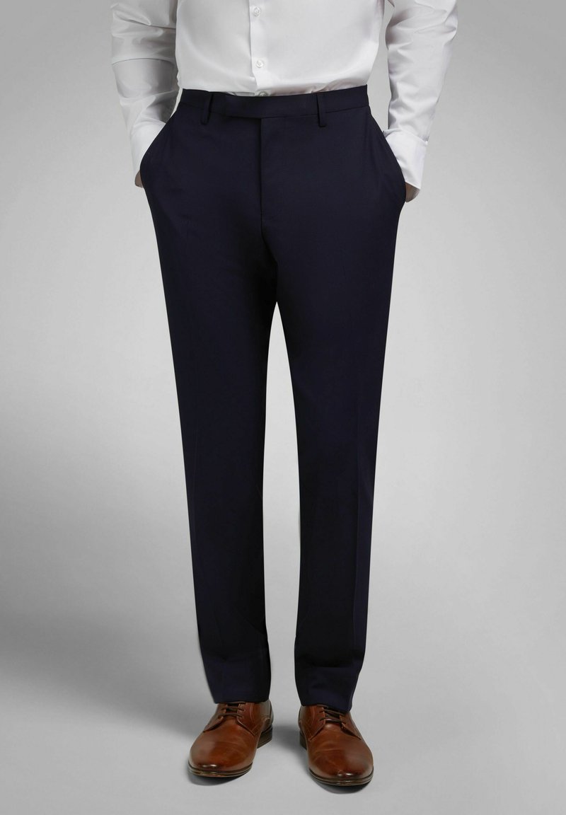 Man draagt marineblauwe nette broek, wit overhemd ingestopt, handen in zijn zakken, en bruine leren nette schoenen tegen een effen grijze achtergrond.