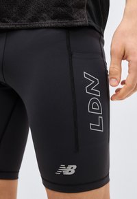 Nærbillede af en person iført sorte træningsshorts med "LDN" og New Balance-logoer på låret og hånden hvilende nær shortsene.