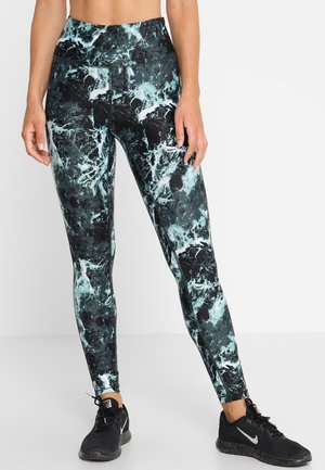Kobieta w czarnych i turkusowych marmurkowych legginsach z wysokim stanem i czarnych butach biegowych Nike stoi na białym tle.