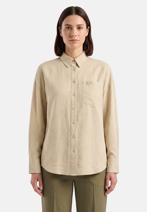 Donna con capelli lisci e scuri che indossa una camicia beige con bottoni e tasca sul petto e pantaloni verde oliva, in piedi su uno sfondo chiaro.