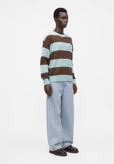 Personne debout portant un pull à manches longues avec de larges rayures marron et bleu clair, un jean large bleu clair et des chaussures marron foncé.
