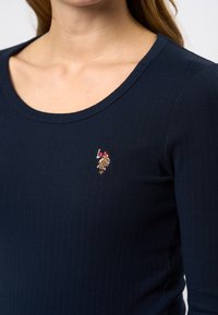 Tumes ribadega topp, millel on madal kaelus, ning millel on vasakus rinnas väike kontrastse värviga broderitud logo.