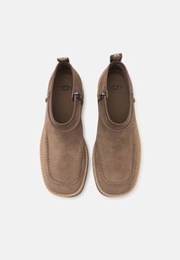 Paire de chaussures marron en daim à enfiler, avec des bouts carrés arrondis, des fermetures éclair latérales et des détails cousus, vues de dessus sur un fond clair.