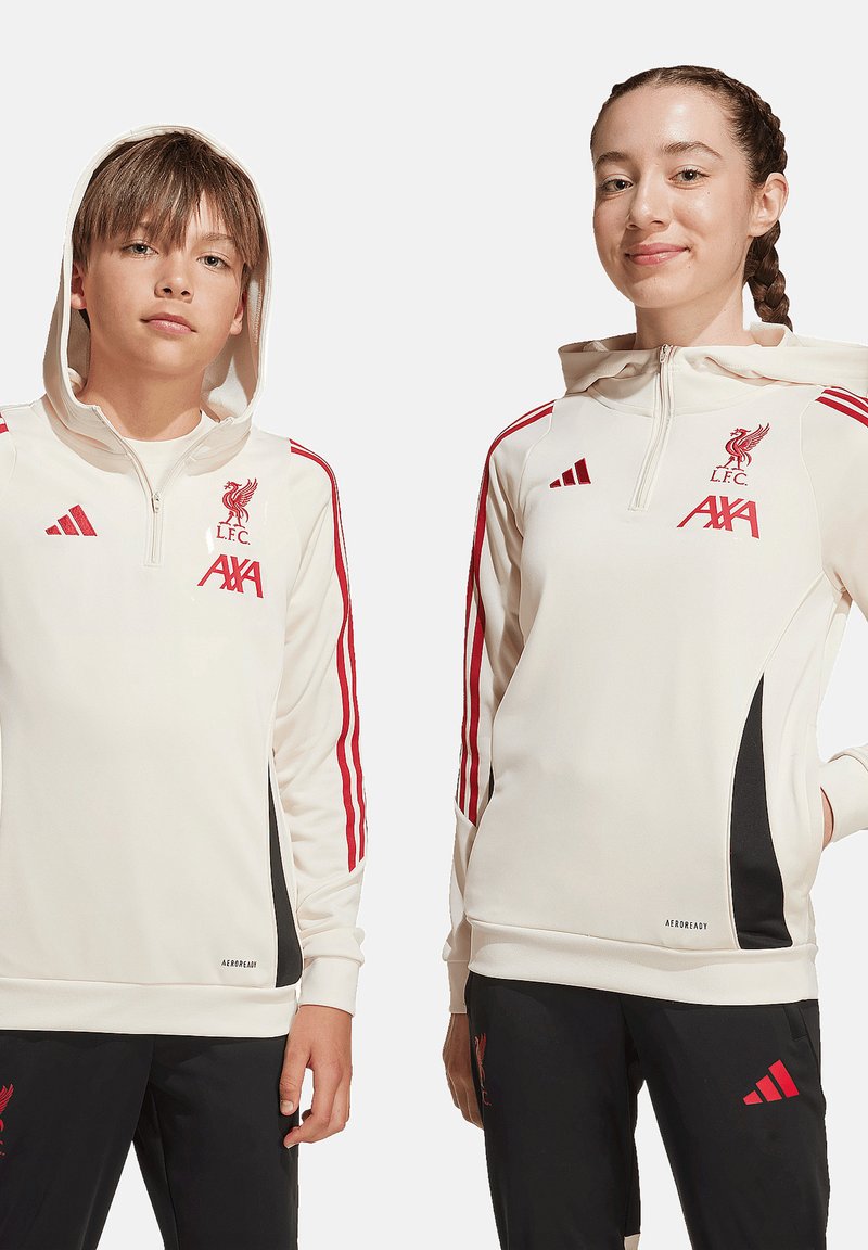 Deux jeunes portent des sweats à capuche blancs du Liverpool FC avec des rayures rouges et un pantalon noir, debout devant un fond uni.