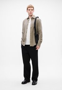 Nuori mies, jolla on päällään beige nappipaita valkoisen T-paidan päällä, mustat löysät housut, mustat loafertossut, kantaen mustaa olkalaukkua valkoista taustaa vasten.