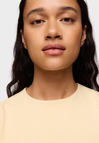 Primer plano de una persona con piel marrón clara que lleva una blusa de cuello redondo de color amarillo pálido. El fondo es sencillo, lo que resalta los rasgos faciales y la joyería sutil.