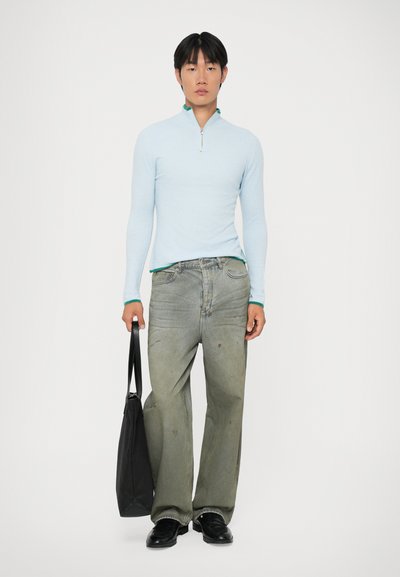Maglietta azzurra a maniche lunghe con colletto con zip, abbinata a jeans verdi sbiaditi a gamba larga e scarpe nere, mentre tiene in mano una borsa a tracolla nera.