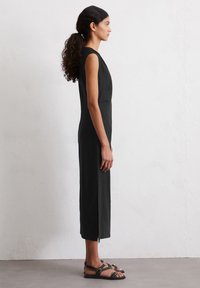 Schwarzes ärmelloses Kleid mit einer figurbetonten Silhouette, knielang, mit einem seitlichen Schlitz. Komplementiert durch flache schwarze Sandalen mit goldenen Akzenten.