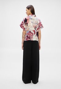 Žena otočená zády, nosící krátký rukáv s abstraktním potiskem a široké černé kalhoty, stojící proti jednoduchému světlému pozadí.