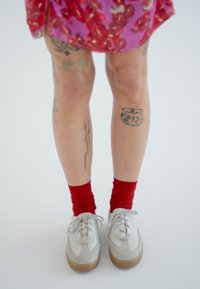 Scarpe low-top bianche e beige con una finitura testurizzata, abbinate a calze a costine rosse. Dettagli dei tatuaggi visibili sulle gambe.