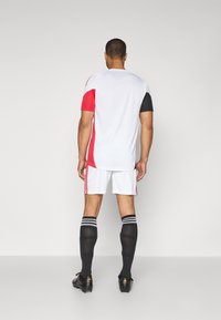 Uniforme de football comprenant un maillot blanc avec des accents rouges et noirs, un short blanc, des chaussettes noires avec des rayures grises et des chaussures noires.