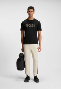 Black T-shirt with "BOSS" logo in gray, cream pants with black side stripes, black sneakers, holding a black backpack.