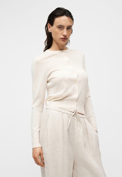 Ανοιχτό μπεζ ζακέτα με στρογγυλή λαιμόκοψη, κούμπωμα με κουμπιά και εφαρμοστά μανίκια, συνδυασμένη με άνετο, ανοιχτόχρωμο λινό παντελόνι.