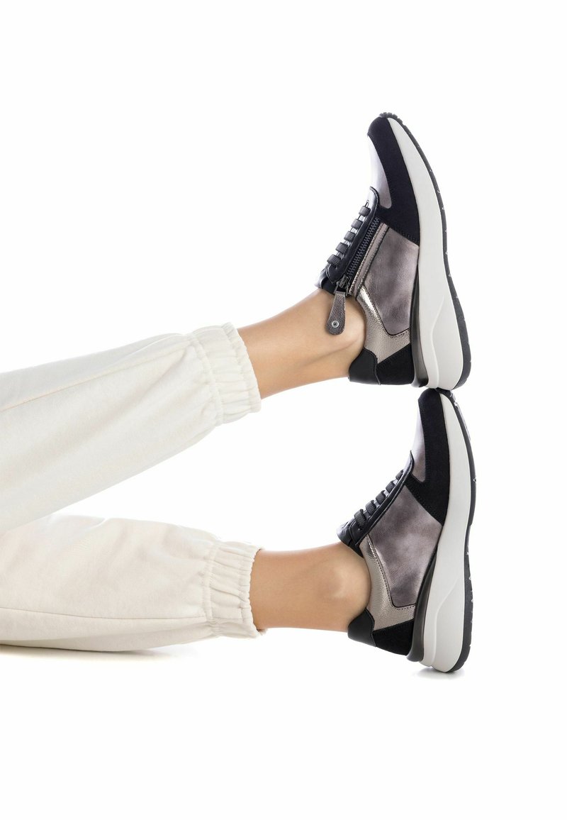 Zapatillas negras y grises con un diseño con cremallera, gamuza texturizada y una suela blanca gruesa, combinadas con pantalones de chándal crema.