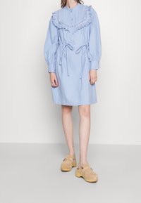 Ljusblå randig klänning med en fransad spetskrage, knappfram, och midjeband. Har långa puffärmar och en knälång skärning.