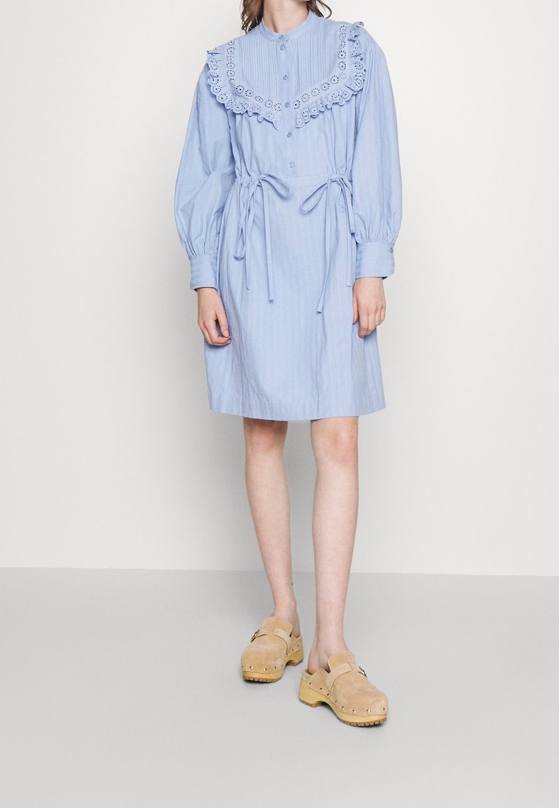 Ljusblå randig klänning med en fransad spetskrage, knappfram, och midjeband. Har långa puffärmar och en knälång skärning.