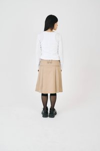 Naine seisab seljaga pöördunud, seljas valge pika varrukaga särk, beež kuni põlveni ulatuv seelik metallrõngastega, mustad sukkpüksid ja mustad platvormkingad.