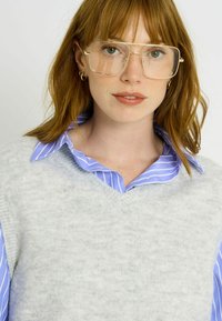 Suéter de punto gris claro con cuello en V, superpuesto sobre una camisa de rayas azules y blancas con cuello. Gafas con montura dorada y pendientes de aro.