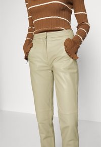 Personne portant un pantalon en cuir beige avec les mains dans les poches et un pull côtelé marron avec des rayures blanches et des poignets à volants.