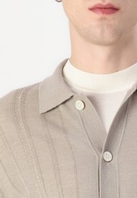 beige πλεκτή ζακέτα με σχεδιασμό γιακά, με υφή ρίγες και δύο κουμπιά στον γιακά, επενδυμένη πάνω από ένα λευκό ριμπ μπλουζάκι.