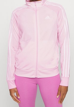 Femme portant une veste de survêtement Adidas rose clair avec des rayures blanches sur les manches et un legging rose assorti, debout devant un fond uni.