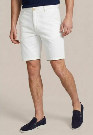 Mann trägt weiße knielange Shorts, marineblaue Slipper und ein dunkelblaues Oberteil vor schlichten Hintergrund.