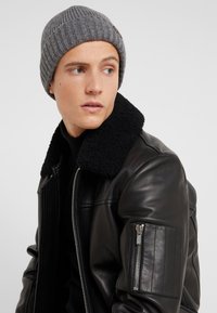 Veste bombardier en cuir noir avec un col en fourrure noire et un bonnet en tricot gris côtelé. Elle dispose d'une poche zippée sur la manche gauche.