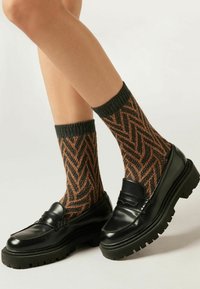 Schwarze Lackleder-Pantoletten mit dicken, gezahnten Sohlen, kombiniert mit braunen und orangen Socken im Fischgrätmuster, die ein geripptes Bündchen haben.
