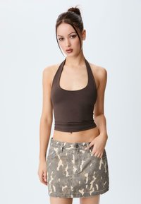 Jeune femme avec des cheveux foncés en chignon, portant un haut en halter marron et une mini-jupe en denim à motif camouflage, se tenant devant un fond blanc uni.