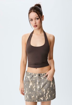Jongere vrouw met donker haar in een knot, gekleed in een bruin haltertop en een denim minirok met camouflagepatroon, staande tegen een effen witte achtergrond.