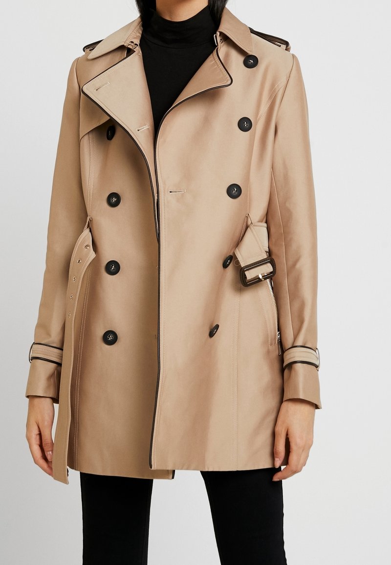 Beige trench coat with black buttons, a double-breasted design, belted waist, and contrasting trim on the collar and cuffs.