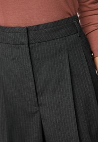 Pantaloni grigi a righe sottili con pieghe, realizzati in tessuto morbido. Vita aderente con dettaglio di chiusura nascosta. Completo abbinato a un top a coste di un rosa tenue.