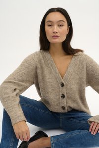 Beige kudumütsiga kardigan sügava V-kaelusega, ees neli tumedat nuppu, soonikvarrukaotsad ja pehme tekstuur. Kantud siniste teksade peal.